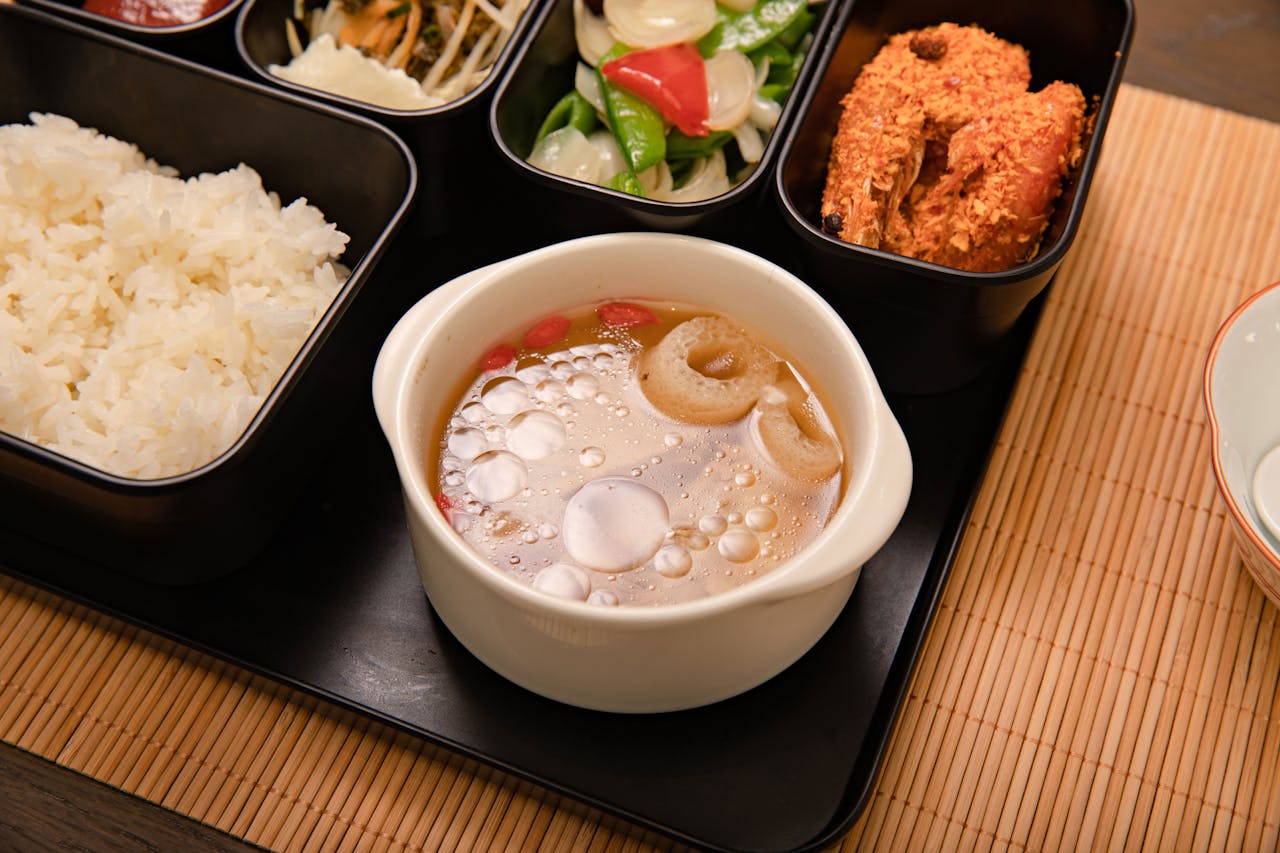 services-06 Asian bento box with rice, fried shrimp, vegetables, and flavorful soup served on a bamboo mat.